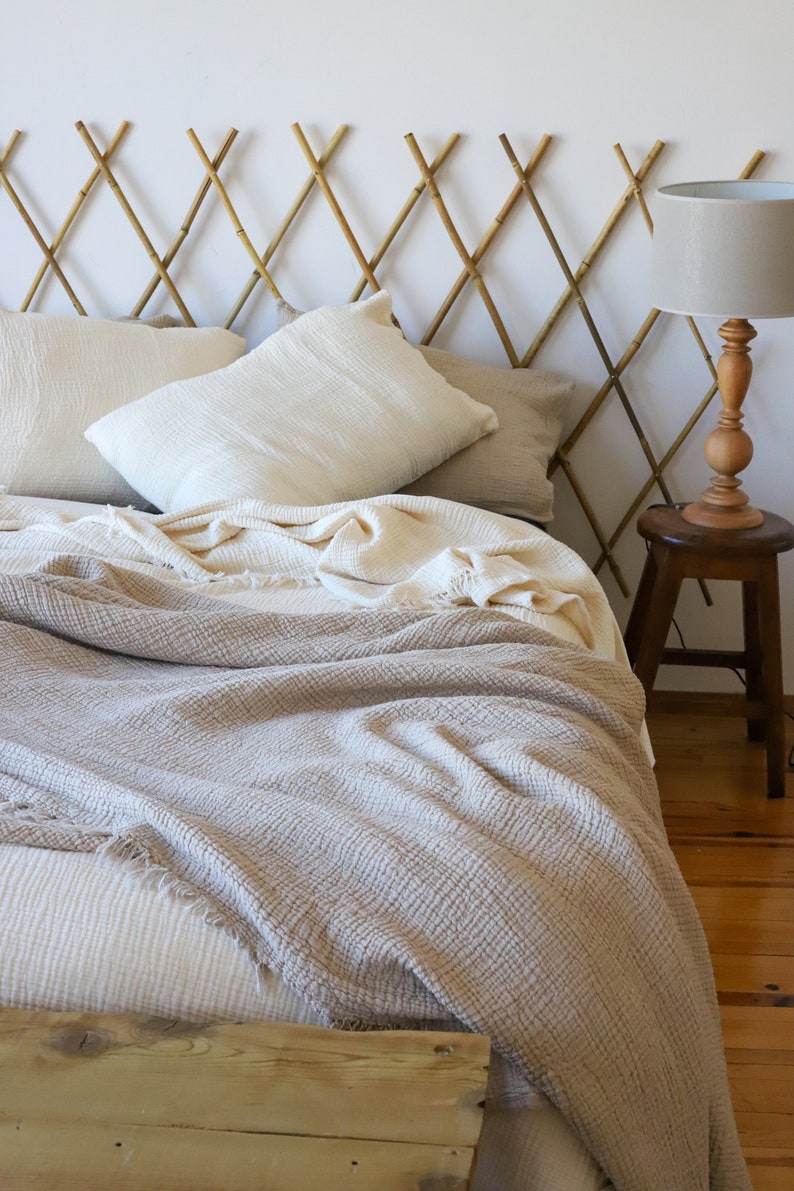 May include: A bed featuring a bamboo headboard, cream-coloured pillows, and a textured blanket in beige and cream hues with a fringed edge. A wooden lamp and stool are positioned beside the bed, creating a cosy bedroom setting.