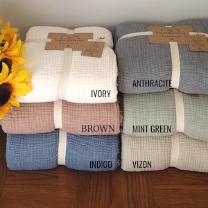 May include: A stack of folded blankets in various colours: ivory, brown, indigo, anthracite, mint green, and vizon. Each blanket is tied with a cream ribbon and a tag. Sunflowers are in the background.