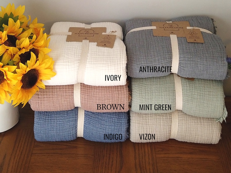 May include: A stack of folded blankets in various colours: ivory, brown, indigo, anthracite, mint green, and vizon. Each blanket is tied with a cream ribbon and a tag. Sunflowers are in the background.
