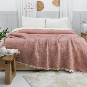 May include: A blush pink waffle weave throw blanket with a fringe border. The blanket is draped over a bed with white pillows and a white bedspread. The blanket is in a bedroom setting with a plant, a wooden bench, and a wicker side table.