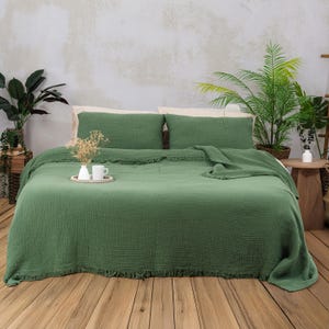 May include: A green bedspread and matching pillowcases are displayed on a bed. The bedspread has a textured appearance and a fringed edge. Two pillows are visible, and a folded blanket rests on the bed. A tray with a cup and vase sits on the bed.