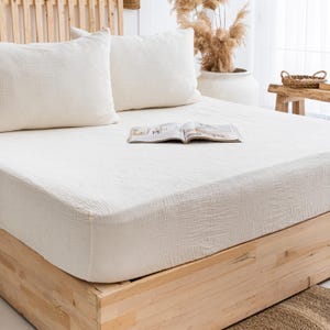May include: A bed with a cream-colored textured fitted sheet and two matching pillows. An open magazine rests on the bed. The bed frame is made of light-colored wood, and the headboard is a wooden slatted design.