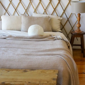 May include: A bedroom with a bed featuring a bamboo headboard. The bed is made with neutral-toned bedding, including pillows and a textured blanket. A wooden bench sits at the foot of the bed, and a wooden side table with a lamp is next to the bed.