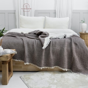 May include: A gray waffle weave throw blanket with a white fringe border. The blanket is draped over a white bed with white pillows. The bed is in a bedroom with a white wall and a plant in a pot on the floor.