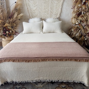May include: A white bed with a brown throw blanket and white pillows. The bed is made with a white duvet cover and has a fringe trim. The bed is in a room with a white wall and a macrame wall hanging.