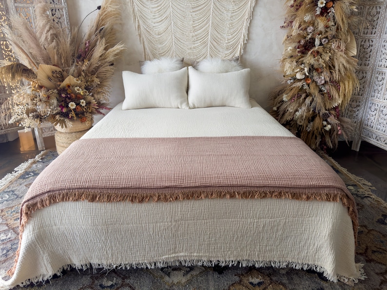 May include: A white bed with a brown throw blanket and white pillows. The bed is made with a white duvet cover and has a fringe trim. The bed is in a room with a white wall and a macrame wall hanging.