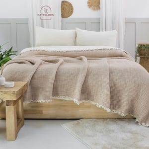 May include: A light brown waffle weave throw blanket with a fringe border is draped over a bed with white linen bedding. The bed is made with two white pillows and a white bolster pillow. The blanket is on a wooden bed frame.
