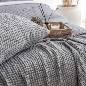 Peut inclure: Un ensemble de literie gris en tissu gaufré, comprenant un oreiller et une couverture, est présenté sur un lit. Un plateau en bois contient une bougie, un diffuseur et des éléments décoratifs. L'ensemble est simple et confortable.