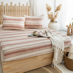 May include: A bed with a light wood frame is covered with a striped comforter and two matching pillowcases. The stripes are a muted red and white. A white fringed throw blanket and a magazine are on the bed. A mug sits on the magazine.