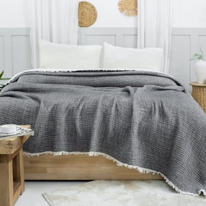 May include: A gray waffle weave throw blanket with a fringed edge is draped over a bed with white pillows. The blanket is made of a soft, textured fabric and has a relaxed, casual look.