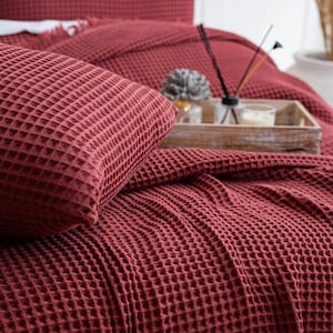 May include: A close-up of a claret waffle-weave bedding set. The set includes a pillow and a blanket, both with a textured, grid-like pattern. A wooden tray with candles and a diffuser sits on the blanket. The overall theme is cozy and inviting.