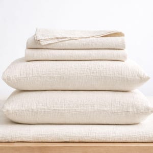 May include: A stack of ivory-colored bedding, including two pillowcases, a fitted sheet, and a folded top sheet. The fabric has a textured, crinkled appearance. The items are neatly arranged, suggesting a focus on comfort and home decor.