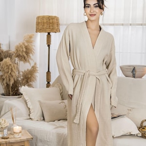 May include: A beige dressing gown with long sleeves and a tie belt. The gown features a V-neck and pockets. The model is wearing the gown in a room with a neutral colour scheme. The robe is made of a soft, textured fabric.