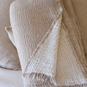 May include: Beige cotton blanket with a textured, crinkled appearance. The blanket has a fringed edge and is folded over pillows, creating a cosy, inviting aesthetic. The neutral colour palette suggests a focus on comfort and relaxation.