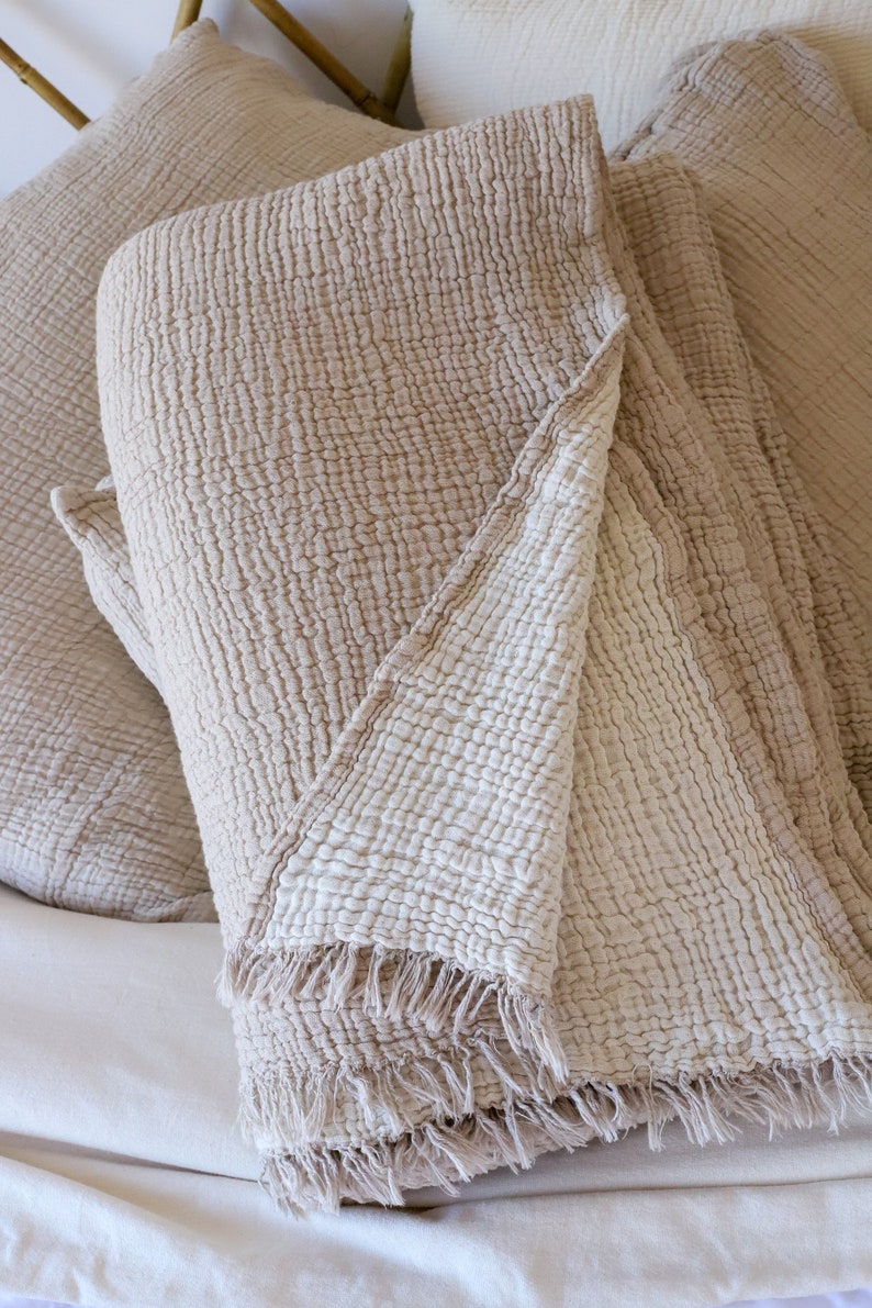 May include: Beige cotton blanket with a textured, crinkled appearance. The blanket has a fringed edge and is folded over pillows, creating a cosy, inviting aesthetic. The neutral colour palette suggests a focus on comfort and relaxation.