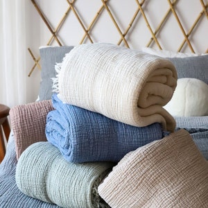 May include: A selection of rolled-up, textured blankets in pastel shades of cream, blue, and pink. The blankets are stacked on a bed with a blue bedspread and a bamboo headboard. The blankets have a soft, inviting appearance.