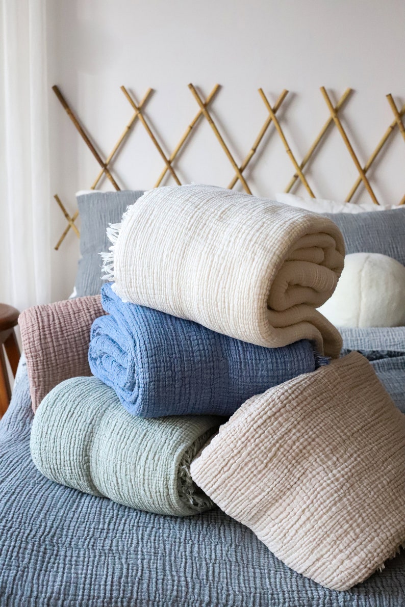 May include: A selection of rolled-up, textured blankets in pastel shades of cream, blue, and pink. The blankets are stacked on a bed with a blue bedspread and a bamboo headboard. The blankets have a soft, inviting appearance.