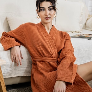 May include: An orange-brown robe with a textured, crinkled appearance. The robe has long sleeves, a wrap-around design, and a matching belt. The model is wearing gold hoop earrings. The robe is made of a soft, comfortable material.