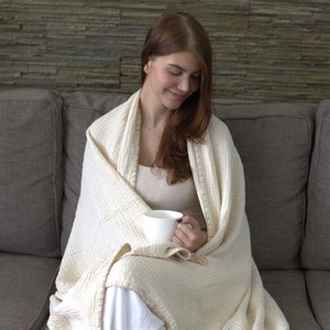 May include: A woman sitting on a couch wrapped in a white waffle weave throw blanket, holding a white ceramic mug.