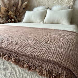 May include: A soft, brown throw blanket with a textured weave and fringe trim. The blanket is folded on a white bed with white pillows.