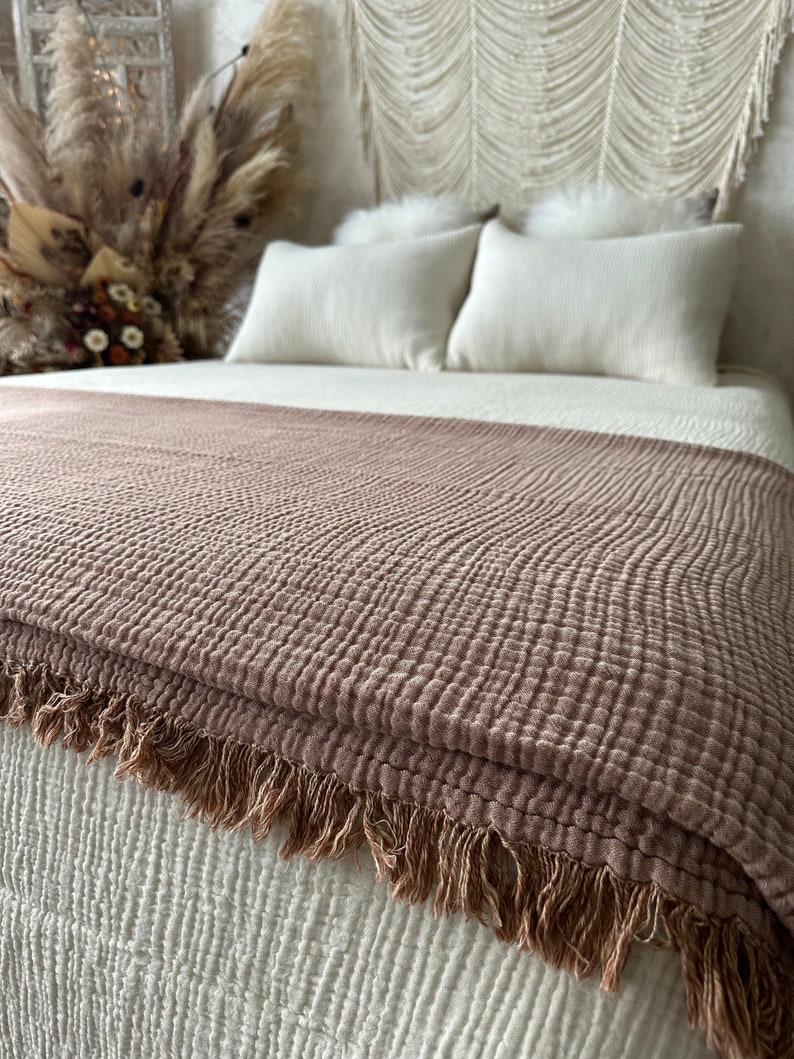 May include: A soft, brown throw blanket with a textured weave and fringe trim. The blanket is folded on a white bed with white pillows.