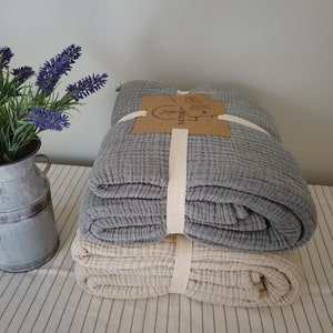 May include: Two folded blankets, one grey and one cream, are stacked and tied with a cream-coloured ribbon. A small metal jug with lavender flowers sits to the left. The blankets appear to be made of a textured fabric.