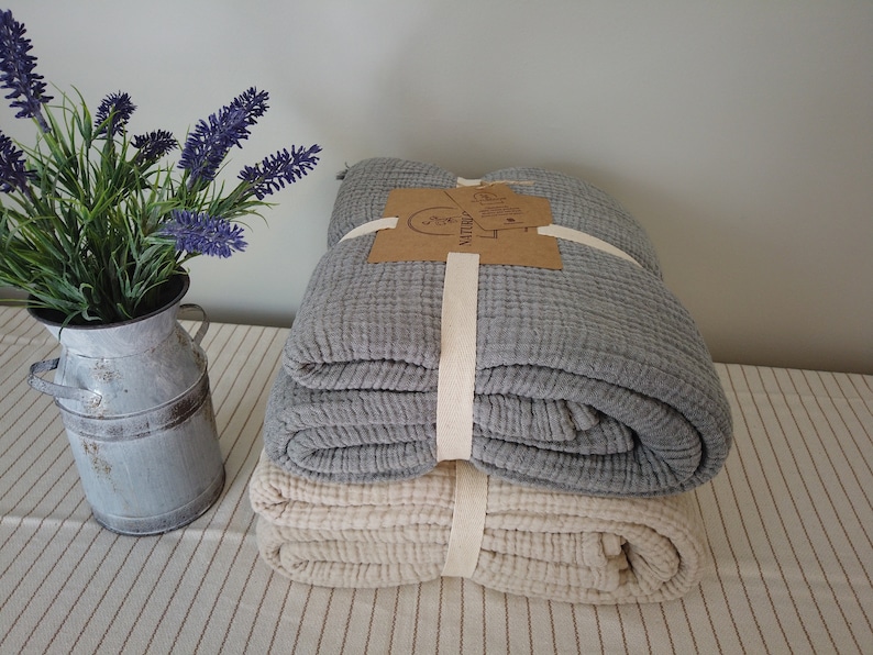 May include: Two folded blankets, one grey and one cream, are stacked and tied with a cream-coloured ribbon. A small metal jug with lavender flowers sits to the left. The blankets appear to be made of a textured fabric.