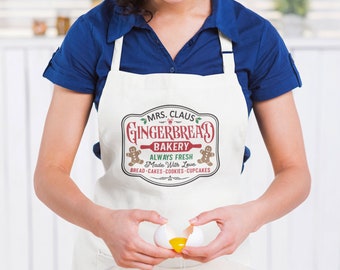 Könnte beinhalten: Eine Person, die eine weiße Schürze mit dem Text "MRS. CLAUS GINGERBREAD BAKERY" trägt, schlägt ein Ei in eine Glasschüssel. Auf der Schürze steht auch "ALWAYS FRESH Made With Love" und listet Backwaren auf. Ein Eierkarton und ein Nudelholz sind sichtbar.
