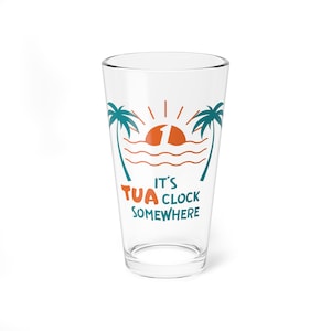 May include: A clear glass with a teal and orange design that says "It's Tua Clock Somewhere". The design features palm trees, a sunset, and waves.