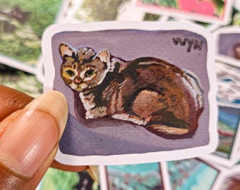May include: A sticker featuring a watercolor painting of a brown tabby cat with green eyes. The cat is lying down on a purple background. The sticker has the text "WYH" in the top right corner.
