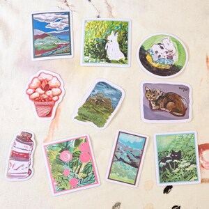 May include: A set of ten colorful stickers featuring various illustrations, including a landscape, a cat, a rabbit, a flower, and a bottle. The stickers are printed on white paper and have a glossy finish.