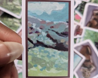May include: A sticker with a painting of a landscape with a blue sky, white clouds, and green and brown hills.