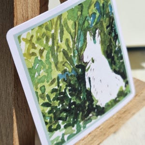 May include: A watercolor painting of a white rabbit peeking out from behind green foliage. The rabbit is facing to the right and is partially obscured by the leaves. The background is a soft green and blue.