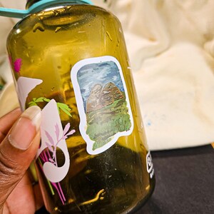 May include: A green, brown, and blue sticker of a mountain range on a brown water bottle. The sticker is slightly peeling at the edges.