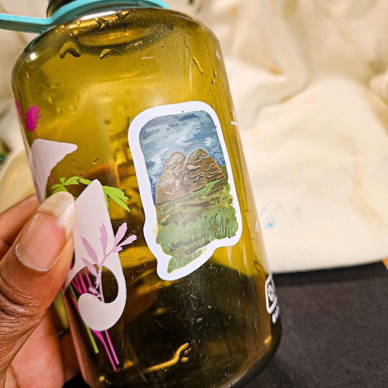 May include: A green, brown, and blue sticker of a mountain range on a brown water bottle. The sticker is slightly peeling at the edges.