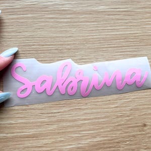 May include: A pink vinyl decal featuring the name Sabrina in a cursive script font, presented on a clear transfer sheet. The lettering is smooth and matte, designed for application onto various surfaces as a personalized decorative label.