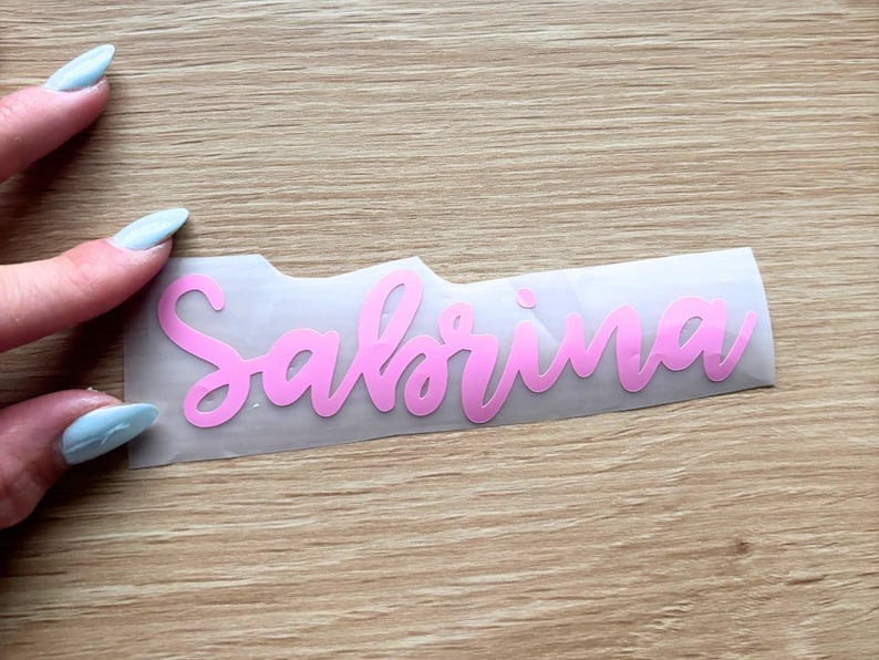 May include: A pink vinyl decal featuring the name Sabrina in a cursive script font, presented on a clear transfer sheet. The lettering is smooth and matte, designed for application onto various surfaces as a personalized decorative label.
