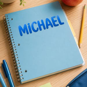 May include: A light blue spiral-bound notebook with the name MICHAEL printed in bold blue letters on the front cover. The notebook features three punched holes along the left side and is positioned alongside two blue pens, a yellow pencil, and a red apple.