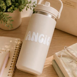 May include: A beige insulated water bottle with a white lid and handle, featuring the name ANGIE in white, retro-style lettering on the front. The bottle has a smooth, matte finish and a metallic rim just below the lid, standing upright.