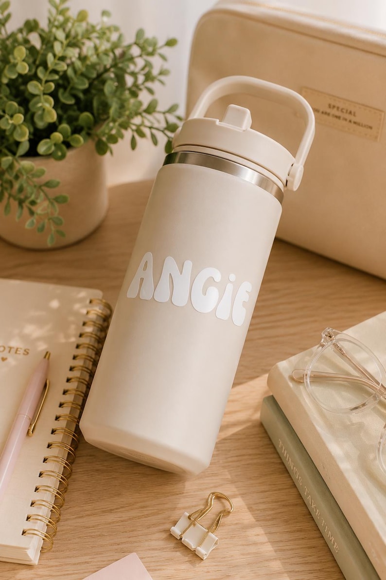 May include: A beige insulated water bottle with a white lid and handle, featuring the name ANGIE in white, retro-style lettering on the front. The bottle has a smooth, matte finish and a metallic rim just below the lid, standing upright.
