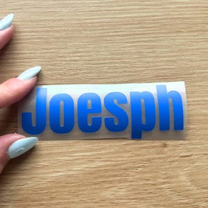 May include: A rectangular adhesive decal featuring the name Joesph in bold, sans-serif blue capital letters. The lettering is printed on a clear, flexible backing material, designed for application onto smooth surfaces as a custom label or sticker.