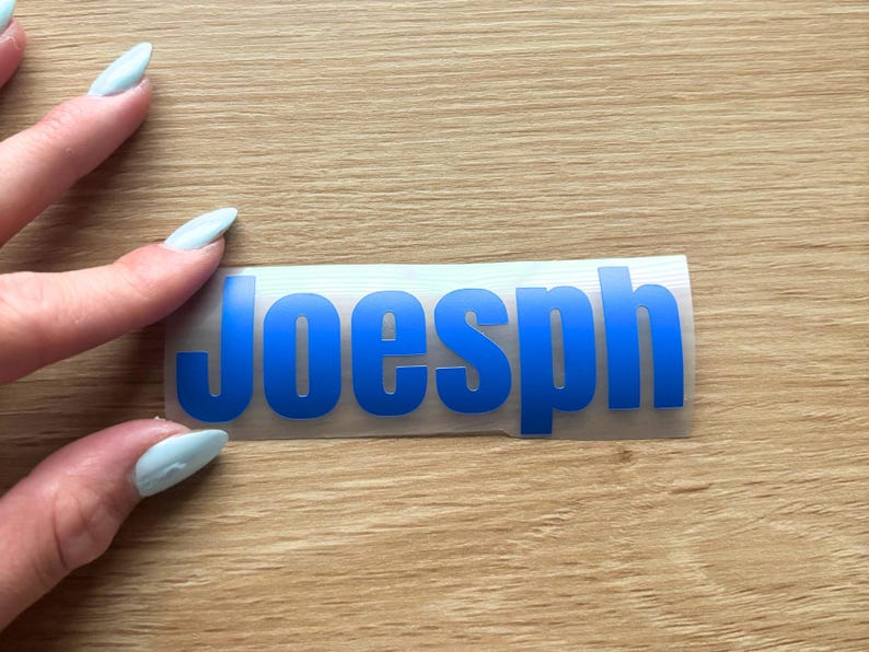 May include: A rectangular adhesive decal featuring the name Joesph in bold, sans-serif blue capital letters. The lettering is printed on a clear, flexible backing material, designed for application onto smooth surfaces as a custom label or sticker.