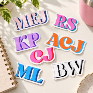 May include: A collection of seven die-cut vinyl stickers featuring two-letter monograms in various color combinations including pink and blue, purple, orange and blue, bright pink, and black and white with a drop shadow effect on each letter.