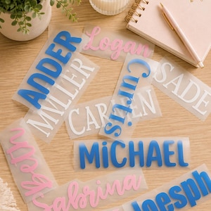 May include: A collection of rectangular clear adhesive labels featuring various names printed in different fonts and colors, including blue, pink, and white. The names visible are Ander, Miller, Logan, Curtis, Sade, Caren, Michael, Beth, Sabrina, Chloe, and Joesph.