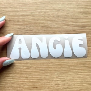 May include: A white vinyl decal featuring the name ANGIE in a bold, rounded, bubble-style font. The letters are cut out and presented on a clear transfer sheet, ready for application to a smooth surface.
