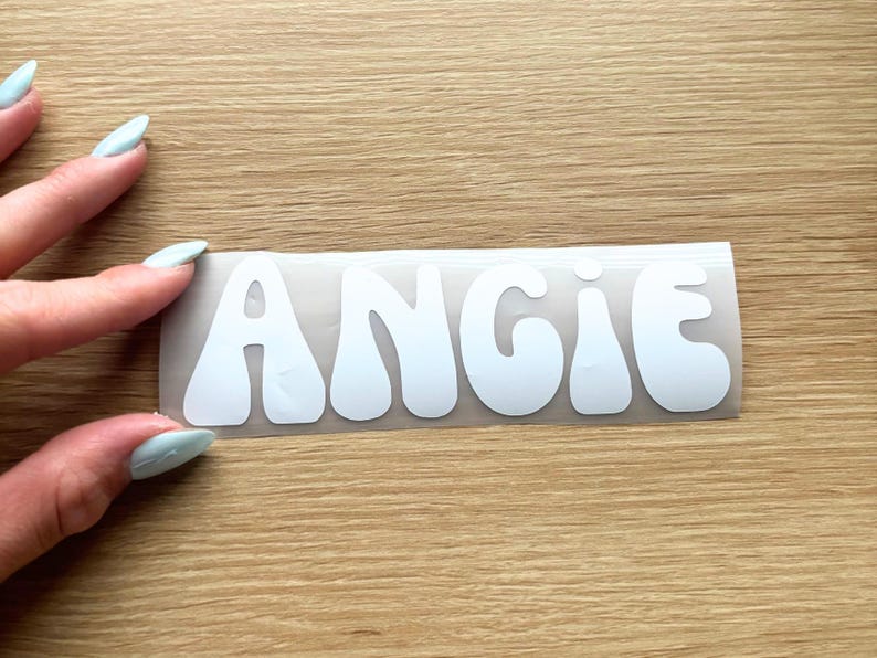 May include: A white vinyl decal featuring the name ANGIE in a bold, rounded, bubble-style font. The letters are cut out and presented on a clear transfer sheet, ready for application to a smooth surface.