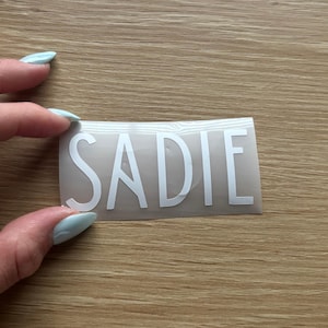 May include: A rectangular adhesive decal featuring the name SADIE in bold, white, sans-serif capital letters. The lettering is applied to a clear, smooth transfer film, creating a clean and simple design suitable for various smooth surfaces.