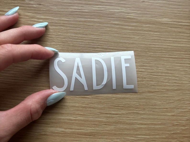 May include: A rectangular adhesive decal featuring the name SADIE in bold, white, sans-serif capital letters. The lettering is applied to a clear, smooth transfer film, creating a clean and simple design suitable for various smooth surfaces.