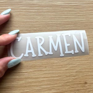 May include: A white vinyl decal featuring the name CARMEN in a bold, sans-serif font. The lettering is presented on a clear transfer sheet, designed for application onto smooth surfaces. The text is clearly legible in uppercase white letters.