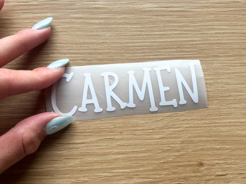 May include: A white vinyl decal featuring the name CARMEN in a bold, sans-serif font. The lettering is presented on a clear transfer sheet, designed for application onto smooth surfaces. The text is clearly legible in uppercase white letters.